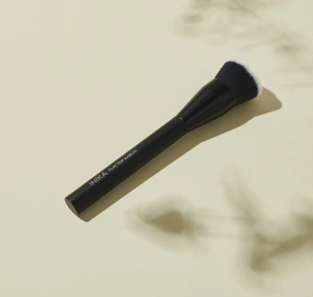 INIKA Organic flat-top brush with an elegant black handle and dense flat bristles, shown on a light beige background.