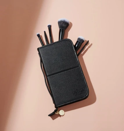 Black makeup brush case with a zipper and gold charm, with different INIKA brushes visible inside on a light peach background.