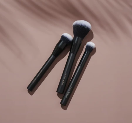 Three INIKA Organic makeup brushes with matte black handles, logo and use labels, and dense gradient bristles lighter at the tips.