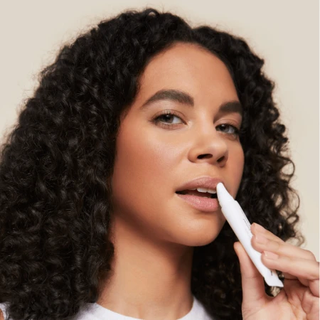 Woman with dark curly hair applying a Jane Iredale lip care product from a white tube, with fresh natural makeup.