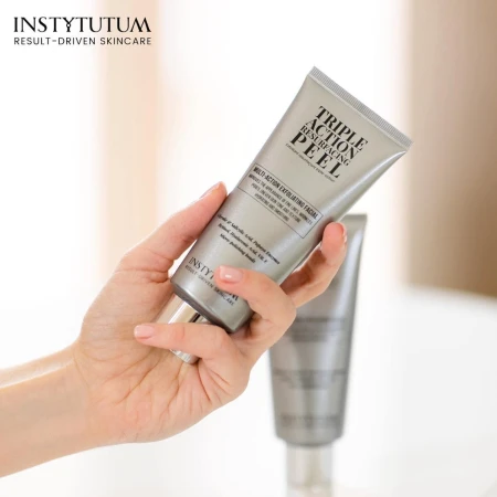Hand holding a silver INSTYTUTUM Triple Action Resurfacing Peel tube with a second tube blurred in the background.