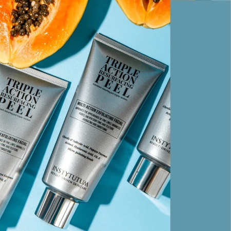 Three INSTYTUTUM Triple Action Resurfacing Peel tubes on a blue background beside a papaya half.
