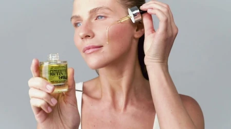 Woman applying INSTYTUTUM Powerful Retin Oil to her cheek with a dropper against a clean grey background.