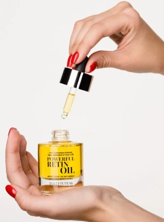 Hands holding INSTYTUTUM Powerful Retin Oil bottle and dropper with yellow oil against a light background.