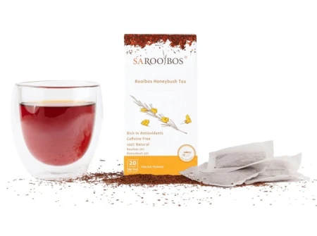 Glass of brewed Rooibos Honeybush tea with SAROOBOS carton, tea bags and loose rooibos leaves beside it.