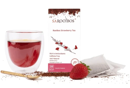 Glass of red Rooibos Strawberry tea with SAROOBOS carton, tea bags, fresh strawberry and golden spoon.