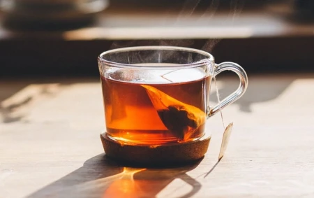 Clear glass of amber rooibos tea on a wooden saucer with a tea bag and visible steam.