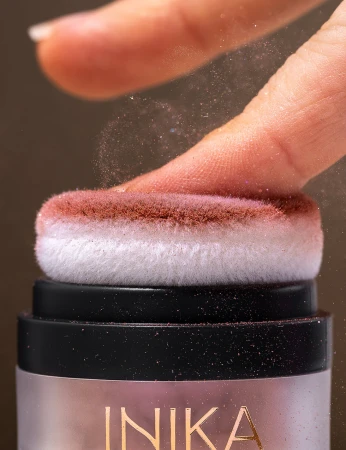 Close-up of the INIKA Organic Puff Pot sponge applicator touching a fingertip with soft rosy powder.