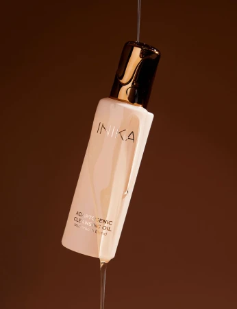 INIKA Organic Adaptogenic Cleansing Oil bottle with a flowing stream of oil against a warm dark background.