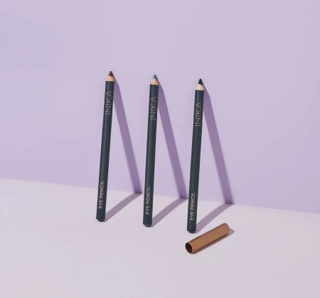 Three INIKA Eye Pencil eyeliners standing upright on a light background with a soft violet tone. Each has a dark barrel and a different colored tip with a gold cap nearby.